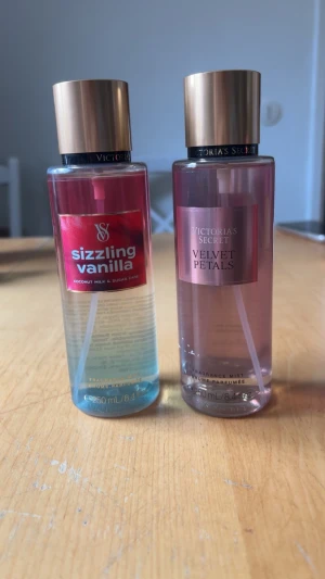 Victoria's Secret bodymist duo - Två Victoria's Secret body mists: Sizzling Vanilla med kokosmjölk och sockerrör i en transparent flaska med rosa och blå toner, samt Velvet Petals med mandel och blomster i en rosa flaska.