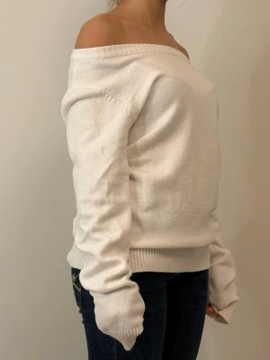 Vit off-shoulder stickad tröja - Snygg vit stickad tröja med off-shoulder design och långa ärmar. Tröjan har ribbade muddar vid ärmslut och nederkant, vilket ger en cool och avslappnad look. Perfekt för dig som gillar att sticka ut med en trendig och bekväm stil.