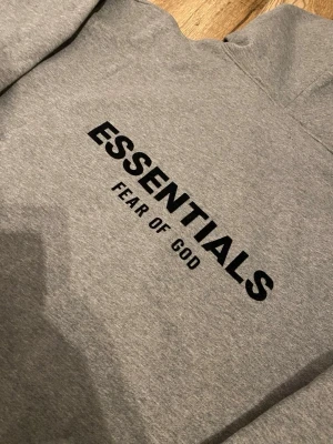 Grå Essentials Fear of God hoodie - Snygg grå hoodie från Fear of God Essentials med svart tryck på ryggen. Klassisk passform med huva och ribbade muddar. Perfekt för dig som gillar streetwear och vill ha en stilren look. Materialet är mjukt och bekvämt, perfekt för vardagsbruk.