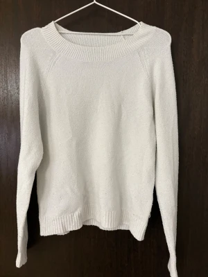 Vit stickad tröja med rund hals - Klassisk vit stickad tröja med rundad halsringning och långa ärmar. Tröjan har ribbade muddar vid ärmslut och nederkant, vilket ger en snygg och enkel look. Perfekt att matcha med jeans eller kjol för en clean stil.