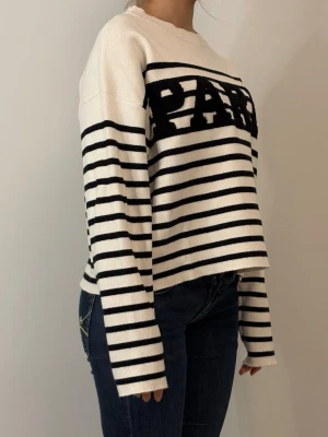 Randig stickad tröja med PARIS-tryck - Vit och svart stickad tröja med breda horisontella ränder och texten PARIS i svart framtill. Tröjan har avslappnad passform, rund halsringning och långa ärmar. Perfekt för en trendig och avslappnad look.