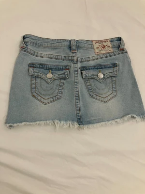 Ljusblå jeanskjol Sadie från True Religion - Snygg ljusblå jeanskjol från True Religion med rå kant nertill och klassiska bakfickor med lock och silverknappar. Kjolen har en kort passform och detaljerade sömmar, perfekt för en trendig look. Storlek 25”