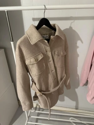 Beige overshirt från NA-KD med bälte - Stilren beige overshirt från NA-KD i mjukt material. Modellen har stora bröstfickor med lock och knappar, bred krage och ett matchande bälte i midjan för en snygg siluett. Perfekt lager-på-lager-plagg med avslappnad passform och långa ärmar.
