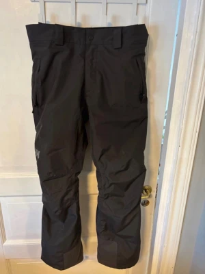 Svarta täckbyxor från Helly Hansen - Säljer ett par svarta täckbyxor från Helly Hansen med raka ben, förstärkta knän och praktiska fickor. Byxorna har bälteshällor och är tillverkade i slitstarkt, vattenavvisande material som passar perfekt för kalla vinterdagar i backen eller på stan. Skicka gärna prisförslag 