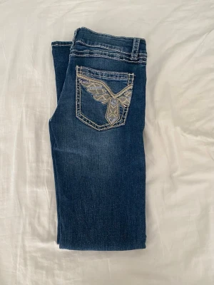 Blå jeans med broderad bakficka - Snygga blå jeans från Gina Tricot med coolt broderi och strass på bakfickan. Jeansen har klassisk femficksdesign, kontrastsömmar och låg midja. Perfekta för dig som vill sticka ut med detaljer och gillar en trendig look.