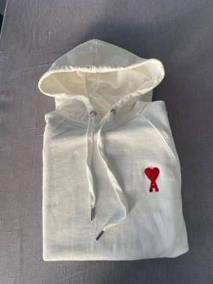 Vit hoodie från Ami med röd logga - Säljer en stilren vit hoodie från Ami med den ikoniska röda hjärtloggan på bröstet. Hoodien har dragsko i huvan, magficka och ribbade muddar. Tillverkad i mjuk bomull för maximal komfort och en clean look. Priset kan diskuteras.
