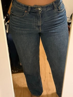 Blå raka jeans med hög midja - Säljer ett par klassiska blå jeans med rak passform och hög midja. Jeansen har fem fickor, bälteshällor och stängs med knapp och dragkedja. Perfekta för en avslappnad och trendig look.