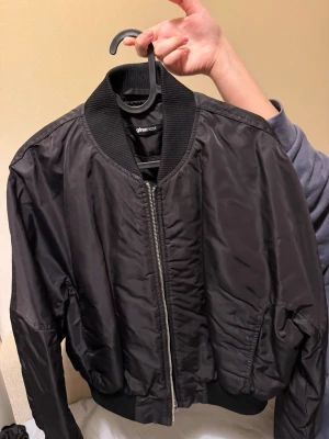 Svart bomberjacka från Gina Tricot - Snygg svart bomberjacka från Gina Tricot med glansig finish och klassisk ribbad krage. Jackan har dragkedja framtill, ribbade muddar vid ärmslut och nederkant samt två fickor på sidorna. Perfekt för en trendig streetstyle-look.