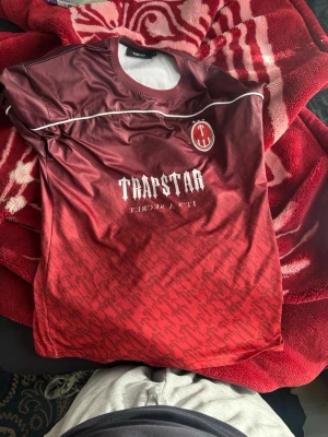 Röd Trapstar t-shirt med tryck - Snygg röd t-shirt från Trapstar med vit logga och text på bröstet samt stort tryck med siffran 22 på ryggen. T-shirten har ett coolt mönster nertill och kontrasterande vita detaljer. Perfekt för dig som vill sticka ut med streetwear-stil.