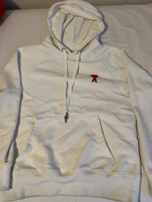 Vit hoodie från Ami Paris med röd logga - Snygg vit hoodie från Ami Paris med klassisk huva och dragsnören. På bröstet sitter den ikoniska röda Ami-loggan med hjärta. Hoodien har en stor magficka och ribbade muddar vid ärmslut och nederkant. Perfekt för en stilren och trendig look.