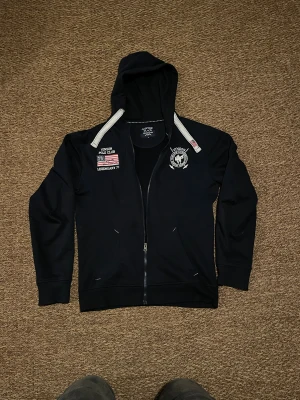 Svart hoodie från Vinson Polo Club - Svart hoodie med dragkedja från Vinson Polo Club. Snygga broderade detaljer med amerikansk flagga och logga på bröstet. Huva med vita snören och två fickor framtill. Perfekt för chill dagar och streetstyle.