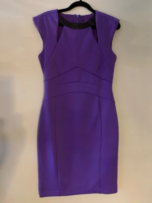 kort klänning - Säljer en snygg lila kortklänning med svarta detaljer vid halsen. Klänningen har en figurnära passform, markerade axlar och är ärmlös. Materialet känns som polyester och dragkedja finns i ryggen. Perfekt för dig som vill sticka ut med färg!