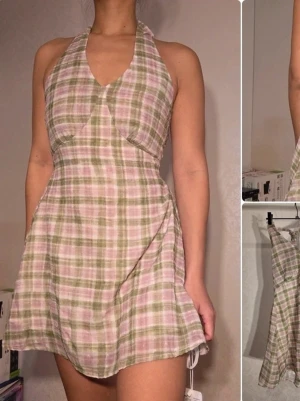 Rutig kortklänning med halterneck från EGGKA - Söt kortklänning med rutigt mönster i ljusrosa, beige och grönt. Klänningen har halterneck, omlott-detalj framtill och är ärmlös. Perfekt för varma dagar och har en lätt, luftig känsla.