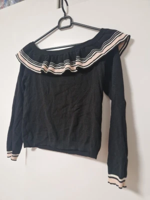 Svart offshoulder topp med volang - Svart offshoulder topp med bred volangkant runt axlarna och vita samt beige ränder. Långärmad modell med matchande randiga muddar vid ärmsluten. 