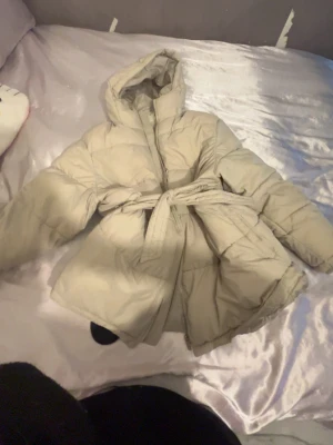 Beige pufferjacka med bälte NLY Trend - Snygg beige pufferjacka från NLY Trend med huva och bälte i midjan. Jackan har quiltad design och dragkedja framtill. Perfekt för kalla vinterdagar och ger en trendig look med sin oversized passform.