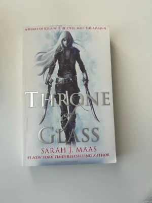 Throne of Glass av Sarah J. Maas - Säljer boken 'Throne of Glass' av Sarah J. Maas. Omslaget är vitt med en fantasyillustration av en kvinnlig karaktär med långt vitt hår och svarta kläder, beväpnad med två svärd. Boken är i pocketformat och tryckt på papper. Perfekt för dig som gillar fantasy och äventyr.