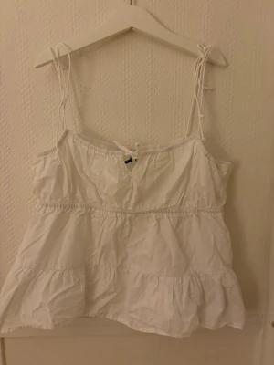 Llinnetopp  - Söt vit linnetopp med tunna axelband som knyts upptill. Toppen har volangiga lager och en luftig, lös passform. Perfekt för varma dagar och enkel att styla med jeans eller kjol.