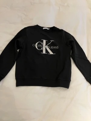 Marinblå  sweatshirt från Calvin Klein storlek 10 - Svart sweatshirt med stor vit CK-logga och texten Calvin Klein Jeans på bröstet. Tröjan har rund hals, långa ärmar och ribbade muddar vid ärmslut och nederkant. Perfekt för en avslappnad och stilren look. Storlek 10 längd axel ner 48 cm armhåla till armhåla 42 cm.