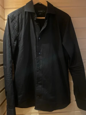 Svart skjorta från Riley - Klassisk svart skjorta från Riley med lång ärm och knappar framtill. Skjortan har en stilren design med normal passform och är tillverkad i ett mjukt bomullstyg. Perfekt för dig som gillar en clean och enkel look.