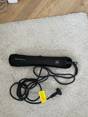 plattång från Björn Axén Tools - En svart plattång från Björn Axén Tools med lång sladd och skyddande fodral. Plattången har en stilren, avlång design och är perfekt för att styla håret. Materialet är plast och metall, och fodralet är i tyg.