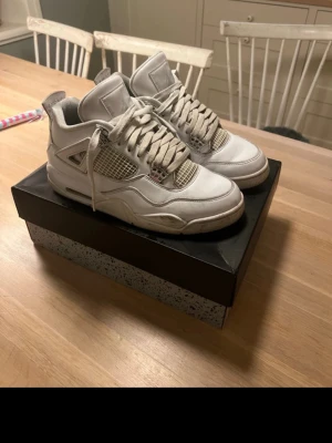 Vita Air Jordan 4 Retro sneakers - Säljer ett par klassiska Air Jordan 4 Retro sneakers i helvitt läder med meshpaneler och ikoniska detaljer. Skorna har tjock sula med synlig Air-enhet och coola plastdetaljer vid snörningen. Perfekta för dig som gillar streetwear och vill sticka ut.