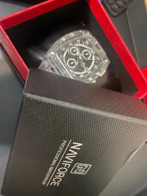 Fyrkantig klocka från Naviforce - Snygg klocka från Naviforce med transparent armband och boett. Klockan har svart urtavla med vita detaljer och tre subdials samt datumvisning. Den fyrkantiga formen och de markerade skruvarna ger en modern och sportig look.