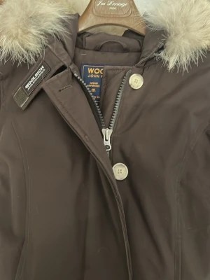 Brun parkas från Woolrich med pälskrage - Snygg brun parkas från Woolrich med stor fluffig pälskrage på huvan. Jackan har dragkedja och knappar framtill samt diskreta sömmar för en clean look. Perfekt för kalla dagar och riktigt klassisk stil.
