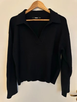 Svart stickad tröja med krage - Säljer en svart stickad tröja från Bik Bok med bred krage och v-ringad hals. Tröjan har lång ärm och en avslappnad passform, perfekt för en stilren och enkel look. Materialet är mjukt och ribbstickat för extra komfort.