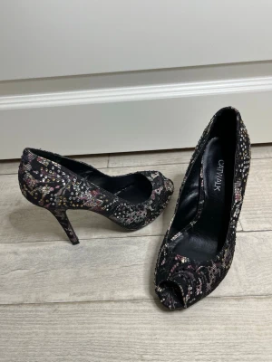 Svarta pumps med paljetter från Catwalk - Snygga svarta pumps i storlek 36 men passar 37 från Catwalk med öppen tå och hög klack. Skorna är täckta av glittriga paljetter i silver, rosa och lila nyanser som ger en festlig look. Materialet är textil med en mjuk insida och klassisk siluett.