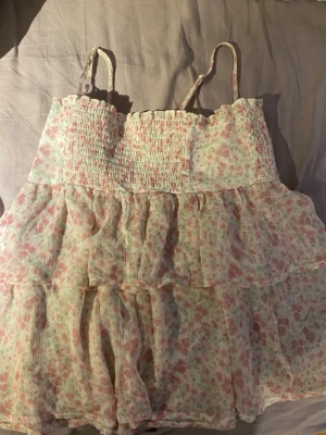 Blommig volangtop med smock - Supersöt blommig linnetopp med smala axelband och smock upptill. Toppen har flera lager volanger och ett ljust, luftigt tyg i rosa och grönt. Perfekt för varma dagar och ger en romantisk vibe.