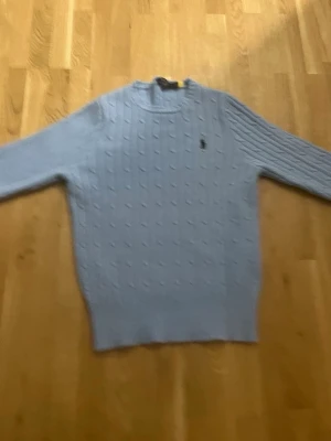 Ljusblå kabelstickad tröja Polo Ralph Lauren - Ljusblå kabelstickad tröja från Polo Ralph Lauren med rund halsringning och klassisk broderad logga på bröstet. Tröjan har långa ärmar och ribbade muddar vid ärmslut och nederkant. Perfekt för dig som gillar en stilren och tidlös look.