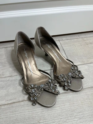 Silvriga pumps med strassdetaljer - Snygga silvriga pumps från Betty Blue i storlek 36 men passar också 37, skorna är med öppen tå och låg klack. Skorna har eleganta strassdetaljer i form av gnistrande rosetter framtill. Materialet är satinliknande och ger en lyxig känsla. Perfekta för dig som vill sticka ut med lite extra bling.