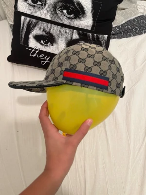 Gucci keps med GG-mönster - Snygg keps från Gucci i grått tyg med klassiskt GG-mönster över hela kepsen. På sidan finns en röd och blå rand som ger en extra touch. Perfekt accessoar för att lyfta din stil.