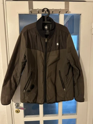 Svart vindjacka från G-Star RAW - Snygg svart vindjacka från G-Star RAW med dragkedja framtill och två fickor med dragkedja. Jackan har en sportig look med diskret logga på bröstet och paneler i olika material. Perfekt för kyliga dagar.