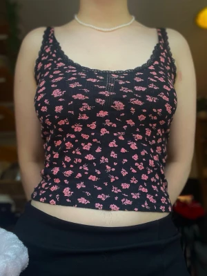 Svart blommig ribbad linnetopp - Supersöt svart ribbad linnetopp med små rosa blommor och spetskant vid urringningen. Toppen är ärmlös och har en figurnära passform som framhäver siluetten. Perfekt att styla med jeans eller kjol för en trendig look. Lappen är avklippt men toppen är från Weekday och passar strl s/m🌸