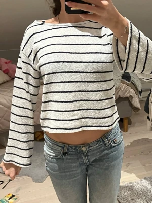 Randig croppad långärmad topp - Vit croppad topp med svarta horisontella ränder och långa ärmar. Toppen har en lös passform och är tillverkad i ett mjukt, frottéliknande material. Perfekt för en avslappnad och trendig look.