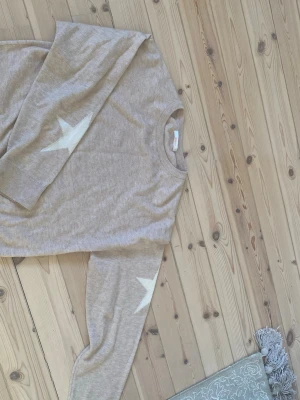 Beige stickad tröja med stjärnor - Snygg beige stickad tröja med rund hals och ribbade muddar.134/149 XS XXS På ärmarna finns vita stjärnor som ger en cool detalj. Tröjan har en avslappnad passform och är perfekt för dig som gillar en enkel men trendig stil. Knappt använd 