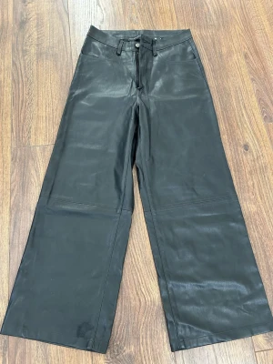 Svarta vida byxor i skinnimitation - Snygga svarta byxor med vida ben och hög midja i skinnimitation. Klassisk femficksmodell med bälteshällor och dragkedja fram. Perfekta för dig som vill ha en edgy och trendig look.