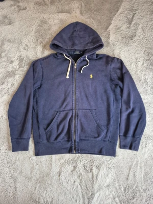 Marinblå hoodie från Polo Ralph Lauren - Snygg marinblå hoodie med dragkedja från Polo Ralph Lauren. Klassisk design med vit dragsko i huvan, kängurufickor och det ikoniska gula Polo-märket broderat på bröstet. Perfekt för en avslappnad och stilren look. Inga hål eller märken