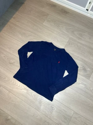 Marinblå långärmad topp från Ralph Lauren - Klassisk marinblå långärmad topp från Ralph Lauren med rund halsringning och den ikoniska röda broderade loggan på bröstet. Tillverkad i mjuk bomull som känns skön mot huden. Perfekt basplagg för en clean och stilren look.