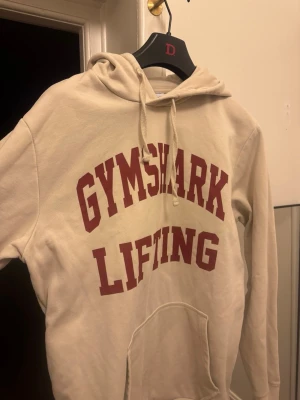 Beige Gymshark hoodie med tryck - Snygg beige hoodie från Gymshark med stort rött tryck 'GYMSHARK LIFTING' på bröstet. Klassisk känguruficka framtill och justerbar huva med snören. Mjuk bomullsmix som känns skön mot huden. Perfekt för gymmet eller chill. Använd max 3 gånger