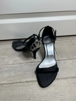 Svarta pumps med strassdetalj - Snygga svarta pumps från Principles i storlek 36 men passar också 37. Dom har en smal klack och öppen tå. Skorna har en elegant rem runt ankeln och en stor strassprydd krona på hälen som ger extra bling. Perfekta för dig som vill sticka ut! Materialet är satinliknande tyg och innersulan är silverfärgad.