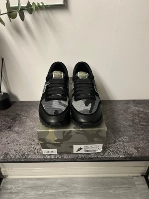 Valentino svart/grå camo sneakers - Säljer ett par snygga Valentino sneakers i svart och grått camomönster. Skorna har mesh- och mockadetaljer, svarta snören och coola nitar på sulan. Perfekta för dig som vill sticka ut med stilrena men ändå unika sneakers.