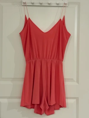 Korallrosa jumpsuit med smala axelband - Snygg korallrosa jumpsuit med smala axelband och öppen rygg. Modellen har resår i midjan för en skön passform och är tillverkad i ett mjukt, lätt tyg som faller fint. Perfekt för varma dagar och enkel att styla med accessoarer.