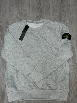 Grå sweatshirt från Stone Island - Snygg gråmelerad sweatshirt från Stone Island med klassisk rund hals och ribbade muddar. Ikonisk svart och gul logotyp-patch på vänster ärm. Perfekt för en clean och avslappnad stil.