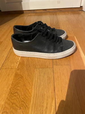 Svarta Arigato sneakers i läder 37 - Snygga svarta sneakers från Axel Arigato i mjukt läder med vita sulor och svarta skosnören. Klassisk låg modell med minimalistisk design och diskret logga på sidan. Perfekta för en clean och stilren look.Storlek 37, innermåttet är 23.5cm.