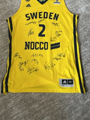 Svensk basketlandslagslinne Adidas signerad av hela landslaget - Ett signerat match linne från svenska basket landslaget - en perfekt vägg prydnad. Märkt med Adidas-logga och sponsorer som NOCCO, SJ och hela landslagets autografer på framsidan. Tillverkad i lätt och ventilerande syntetmaterial, perfekt för basket eller samling. Jag vann den i en match mot Sverige och Tyskland. Oklart om den var med i matchen eller inte. 