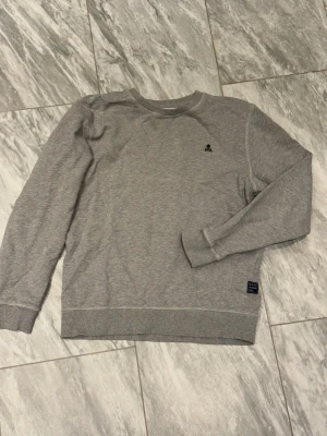 Grå sweatshirt med liten logga - Säljer en stilren grå sweatshirt med rund halsringning och liten broderad logga på bröstet. Tröjan har ribbade muddar vid ärmslut och nederkant, perfekt för en avslappnad look. Materialet är mjukt och bekvämt bomullstyg.