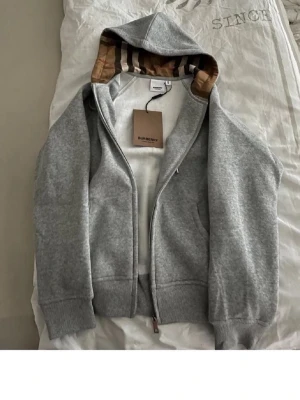 Grå zip hoodie från Burberry - Snygg grå zip hoodie från Burberry med klassisk logga på bröstet och ikoniskt rutigt foder i huvan. Tillverkad i mjuk bomullsblandning, har ribbade muddar och fickor på sidorna. Perfekt för dig som vill ha en stilren och exklusiv look.