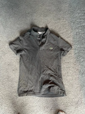 Mörkgrå pikétröja från Lacoste - Säljer en mörkgrå pikétröja från Lacoste med klassisk krage och knappar framtill. Tröjan har korta ärmar och den ikoniska gröna krokodilloggan på bröstet. Perfekt till jeans eller shorts för en clean och enkel look.